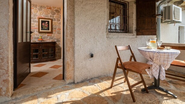 Habitación rústica con pared de piedra, puerta de madera, una mesita con silla y una ventana con barrotes.