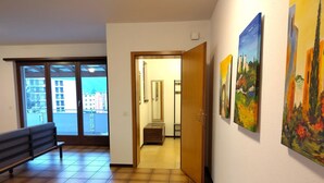 Een gang met een bankje, een deur naar een kamer en uitzicht op gebouwen.