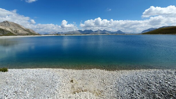 Un lac paisible aux eaux limpides, entouré de reliefs rocheux et bordé par des montagnes lointaines, le tout sous un ciel bleu.