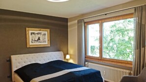 Un dormitorio con cama, una silla, ventana con cortinas, radiador y un cuadro en la pared.
