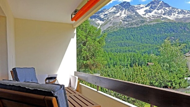 Un balcón con mesa y sillas de madera, con vistas a un valle boscoso y montañas nevadas.