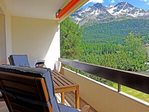 Een balkon met een houten tafel en stoelen, met uitzicht op een bebost dal en besneeuwde bergen.