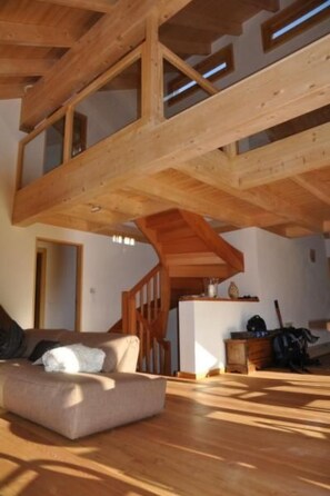 Un loft spacieux avec de hauts plafonds, des poutres apparentes et une grande fenêtre laissant entrer la lumière naturelle. Un canapé est placé près d’une porte, et une petite table avec quelques objets est visible au fond.