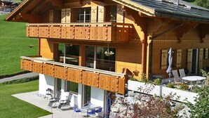 En stuga byggd av trä med en balkong, stora fönster och utsikt över ett grönskande landskap.