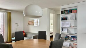 Een moderne woonkamer met een ronde houten tafel, grijs gestoffeerde stoelen, een witte boekenkast en een grote witte hanglamp.