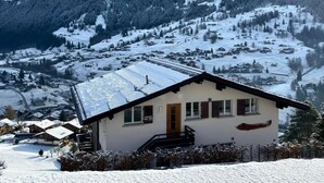 Een bergdorp bedekt met sneeuw, met een klein wit gebouw met een houten deur en ramen, omringd door besneeuwde velden en bomen.