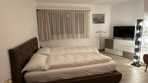 Un dormitorio con cama, mesita de noche, televisor y ventana con cortinas.