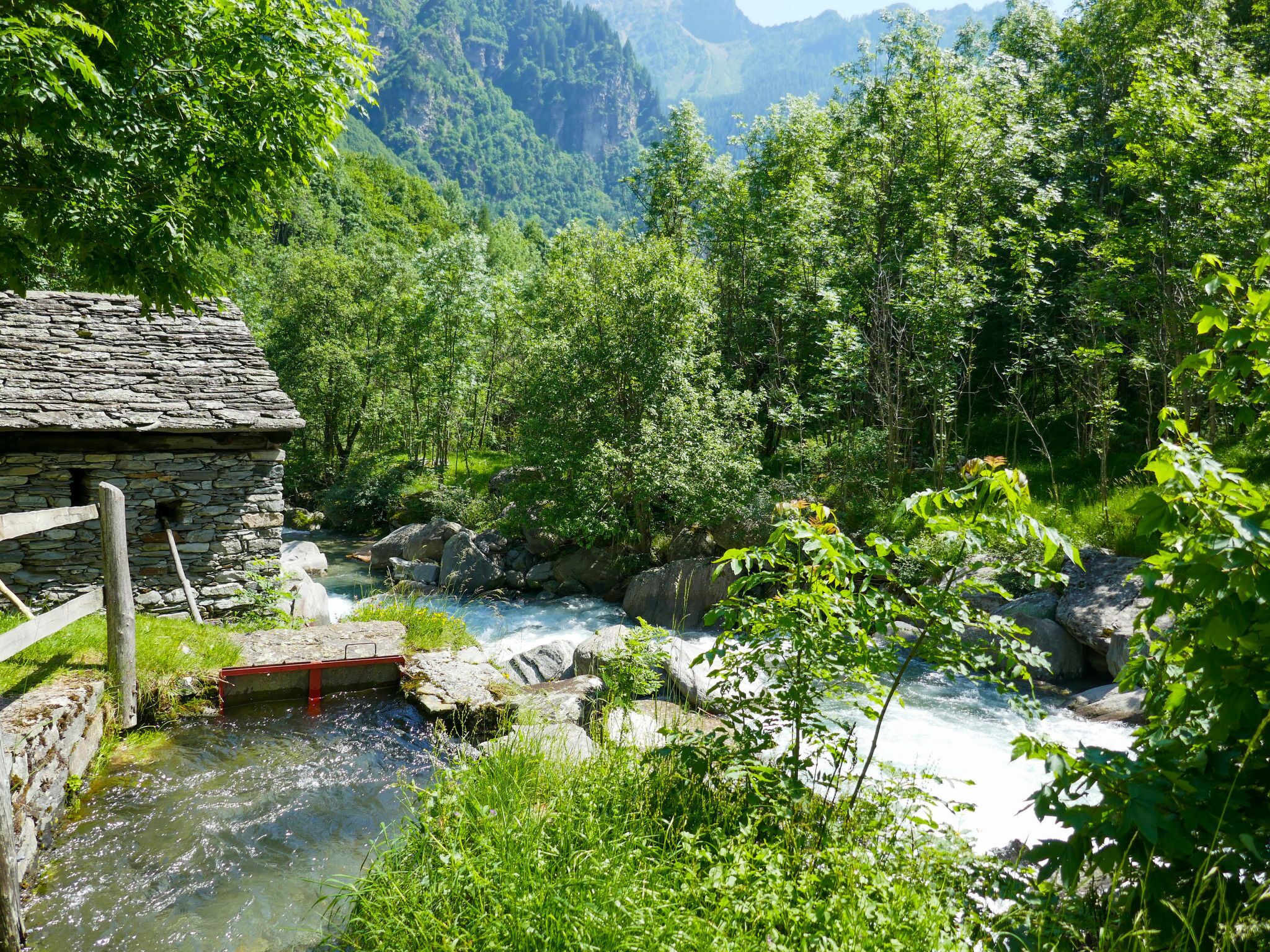 Un edificio rústico de piedra con cerca de madera, un pequeño puente sobre un arroyo cristalino, rodeado de vegetación exuberante y montañas al fondo.