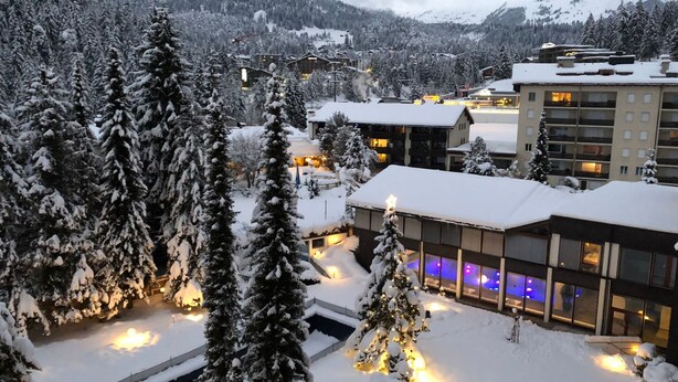Een met sneeuw bedekt wintersportresort met moderne gebouwen, een zwembadgedeelte met verlichte lampen, en een bergachtige achtergrond.