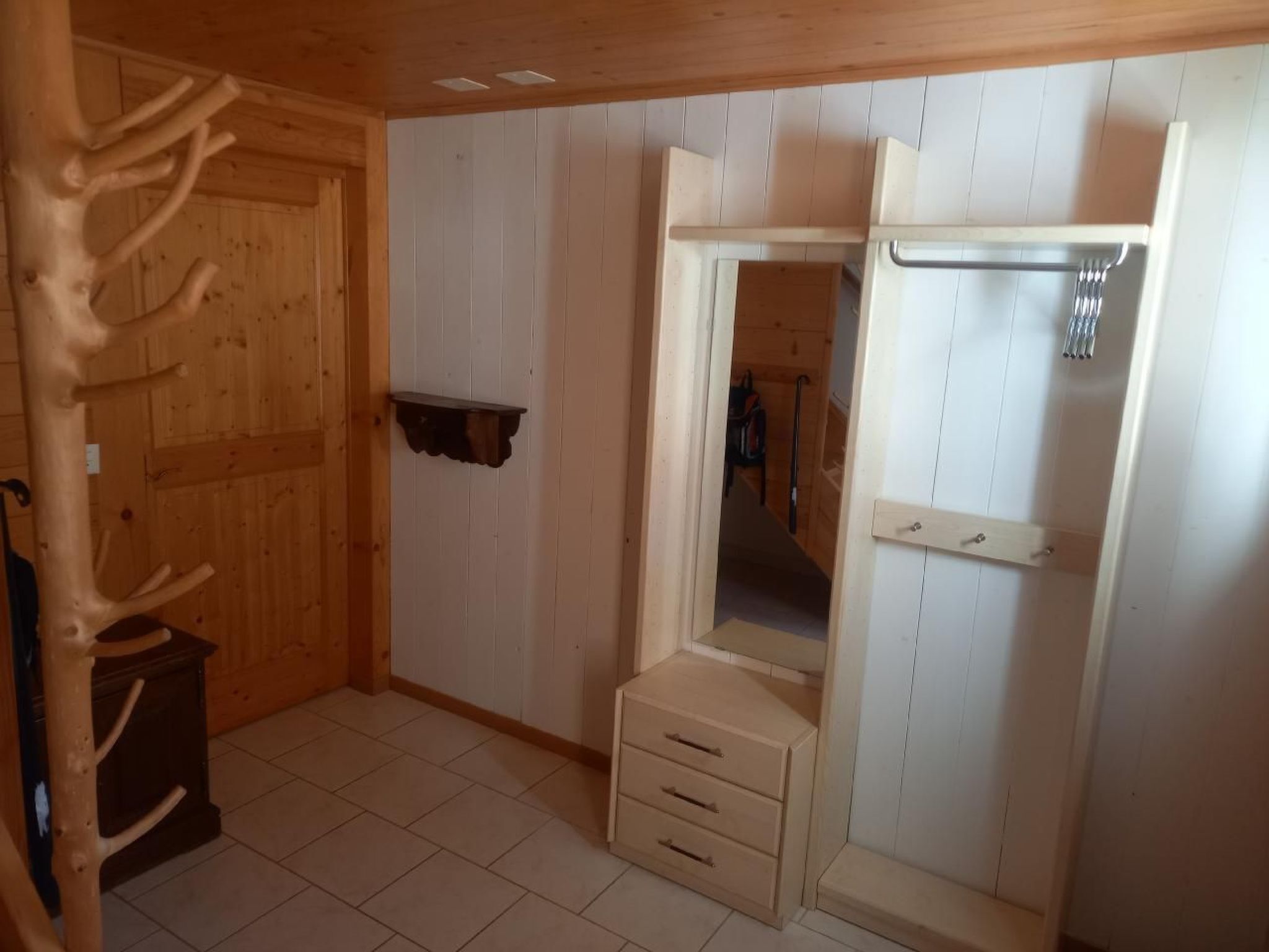 A room with wooden walls and a door, a small cabinet, and a mirror with a shelf above it.