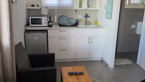 Une cuisine compacte équipée d’un four à micro-ondes, d’un petit réfrigérateur et d’un évier. On y trouve une table en bois sur laquelle est posée une télécommande, ainsi qu’un canapé en osier dans l’espace salon.