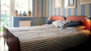 Un dormitorio con paredes a rayas, una cama con ropa de cama a rayas, dos cuadros enmarcados, una ventana con persianas y un radiador.