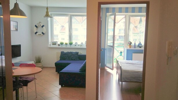 Un salón acogedor con un sofá, una mesita y una televisión. Hay un balcón con vistas a edificios y una puerta que da a otra habitación.