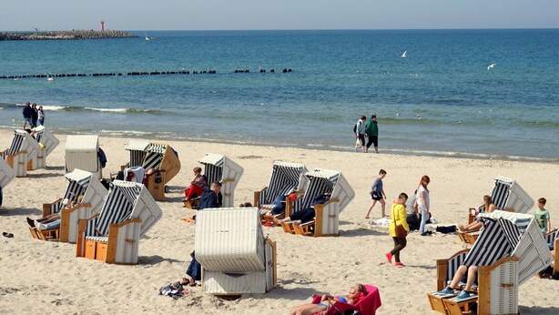 Una playa con hileras de sillas y sombrillas a rayas, un muelle al fondo, y gente caminando por la orilla.