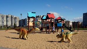 Un parque infantil con temática de dinosaurios, con estructuras para escalar y toboganes, frente a edificios residenciales.