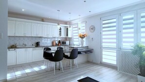 Una cocina moderna con armarios blancos, una zona de comedor con sillas negras y un ventanal con contraventanas.