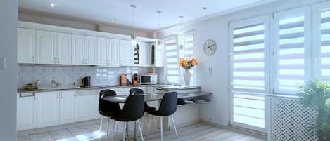 Una cocina moderna con armarios blancos, una zona de comedor con sillas negras y un ventanal con contraventanas.
