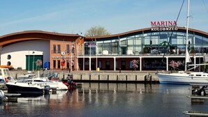 Una marina con barcos amarrados, un edificio con el letrero 'MARINA KOLOBRZEG' y una oficina de 'BIURO PORTOWE'.