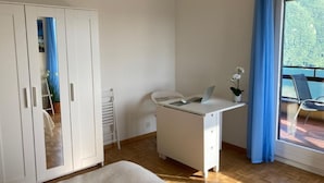 Ein Zimmer mit weißem Schreibtisch, einem Stuhl, einem Kleiderschrank und einem Balkon mit blauen Vorhängen.