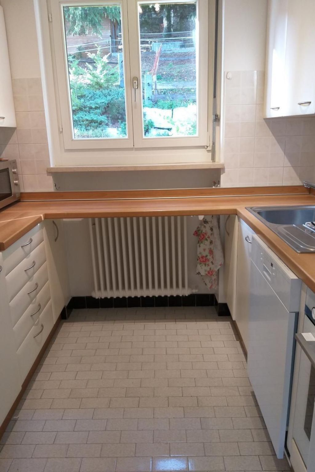 A compact kitchen with a window, white cabinets, a wooden countertop, a radiator, and a dishwasher.