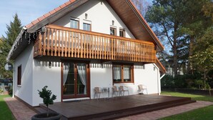 Een huis met twee verdiepingen, met een houten balkon, een klein terras met stoelen en een plant in een pot.