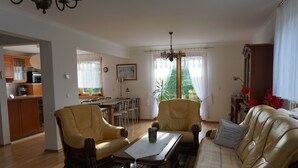 Een woonkamer met een bank, twee fauteuils, een salontafel en een eethoek met een tafel en stoelen. In de achtergrond is een keuken te zien.