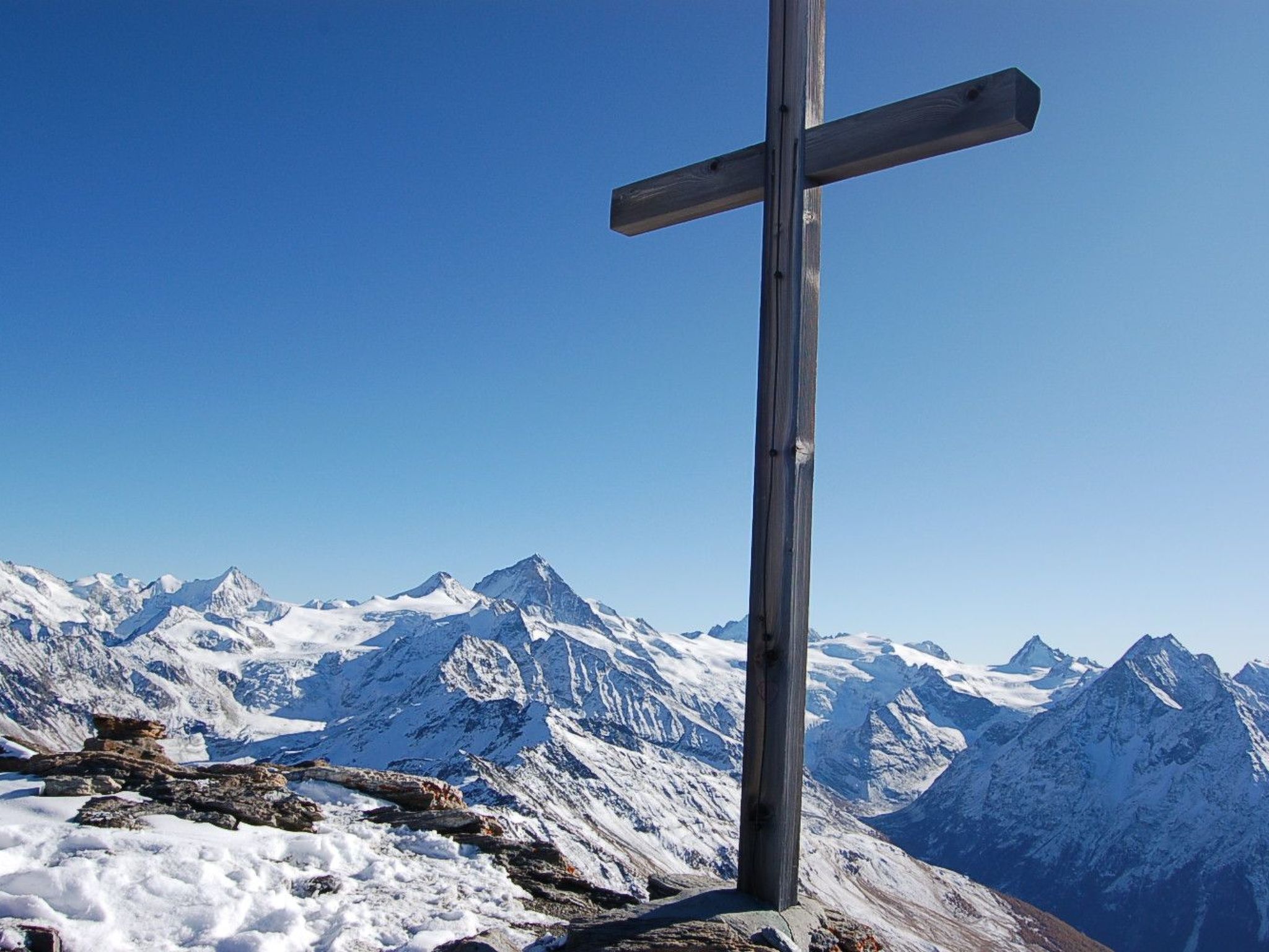 Une croix en bois, dressée au sommet d’une montagne, avec en arrière-plan des pics enneigés.