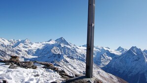 Une croix en bois, dressée au sommet d’une montagne, avec en arrière-plan des pics enneigés.