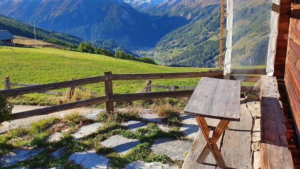 Une cabane en bois, avec un sentier de pierres menant à une table en bois, d’où l’on peut admirer un paysage montagneux sous un ciel dégagé.