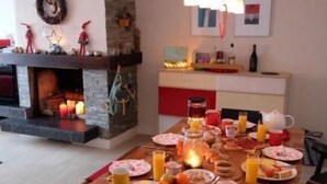 Un coin repas avec une longue table en bois dressée pour le petit-déjeuner, une cheminée et, au fond, une cuisine moderne.