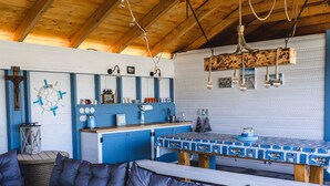 Ein gemütliches Blockhaus-Interieur mit Holzbalkendecke, einem Esstisch mit blau-weiß kariertem Tischläufer und einer blauen Küchenarbeitsplatte.
