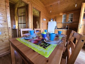 A wooden cabin interior with a dining table set for four, equipped with blue cups and plates, and a refrigerator in the background. - with communal pool by Interhome (Rusinowo)
