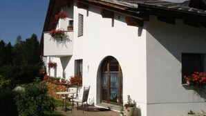 Una casa de dues plantes amb un balcó, un petit pati amb taula i cadires, i un jardí amb gespa i plantes en testos.