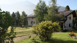 Paisatge rural amb una casa envoltada de verdura, cel blau i muntanyes al fons.