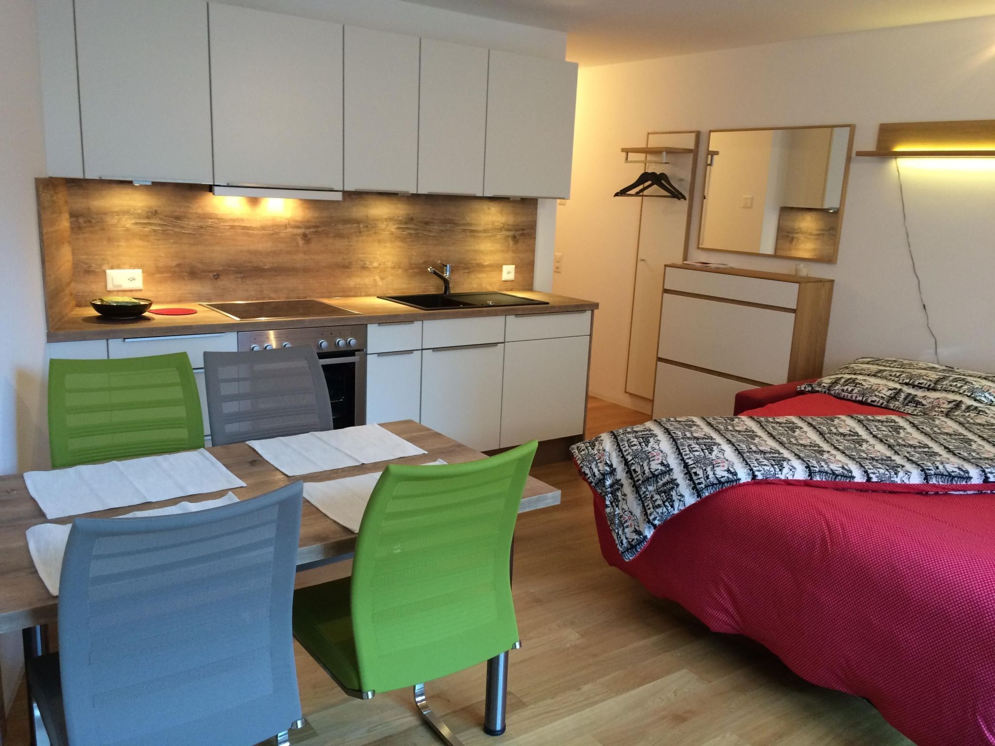 A compact room with a kitchenette, a dining area with a table and chairs, and a bed with a patterned blanket.