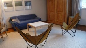 Ein Wohnzimmer mit einem blauen Sofa, einem weißen Couchtisch und zwei modernen Sesseln. Gegen die Wand lehnt eine Holzkommode und an der Wand hängen drei gerahmte Bilder.