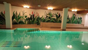Une piscine intérieure aux murs et au plafond recouverts de carreaux, entourée d’un mur de briques orné d’une fresque représentant des plantes.