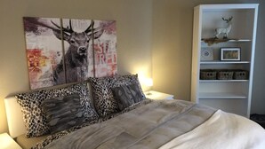 Une chambre à coucher avec un grand lit, une tête de lit ornée d’un tableau représentant un cerf, une étagère blanche agr émentée d’objets décoratifs et une lampe sur la table de chevet.