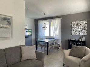 A modern living room with a grey sofa, a dining area with a glass table, and a kitchen with a built-in oven. - Casa Bijou by Interhome (Locarno)
