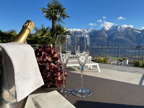 Et bord med en champagneflaske, to vinglas og en blomsterbuket på en terrasse med udsigt til et bjerg.