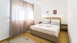 Un dormitorio con cama, cortinas, una mesita de noche, una lámpara de pared y dos cuadros enmarcados.