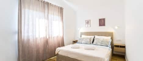 A bedroom with a bed, curtains, a nightstand, a wall-mounted light, and two framed pictures.