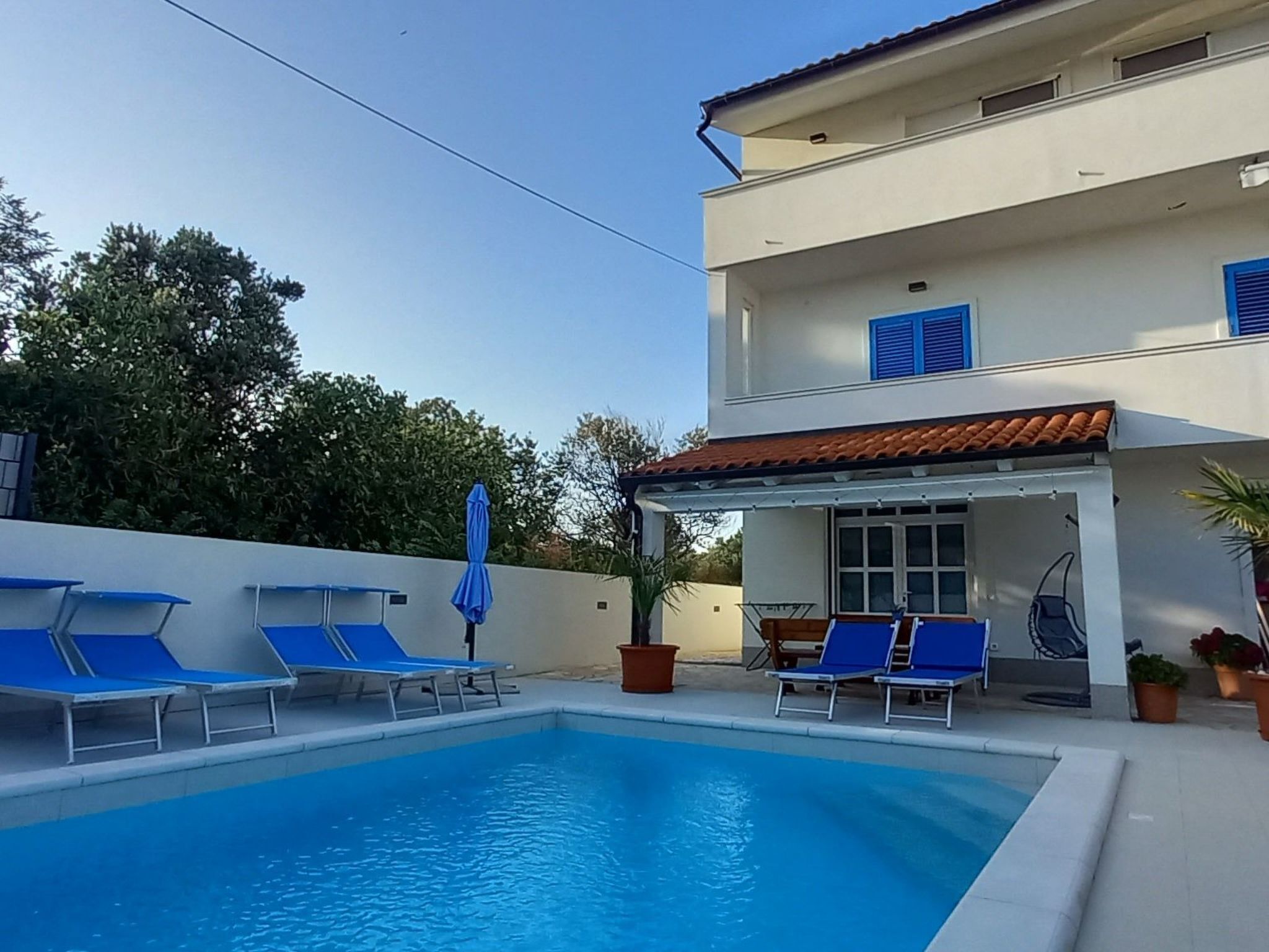 A swimming pool with blue lounge chairs, a covered patio area, and a two-story building with blue shutters.
