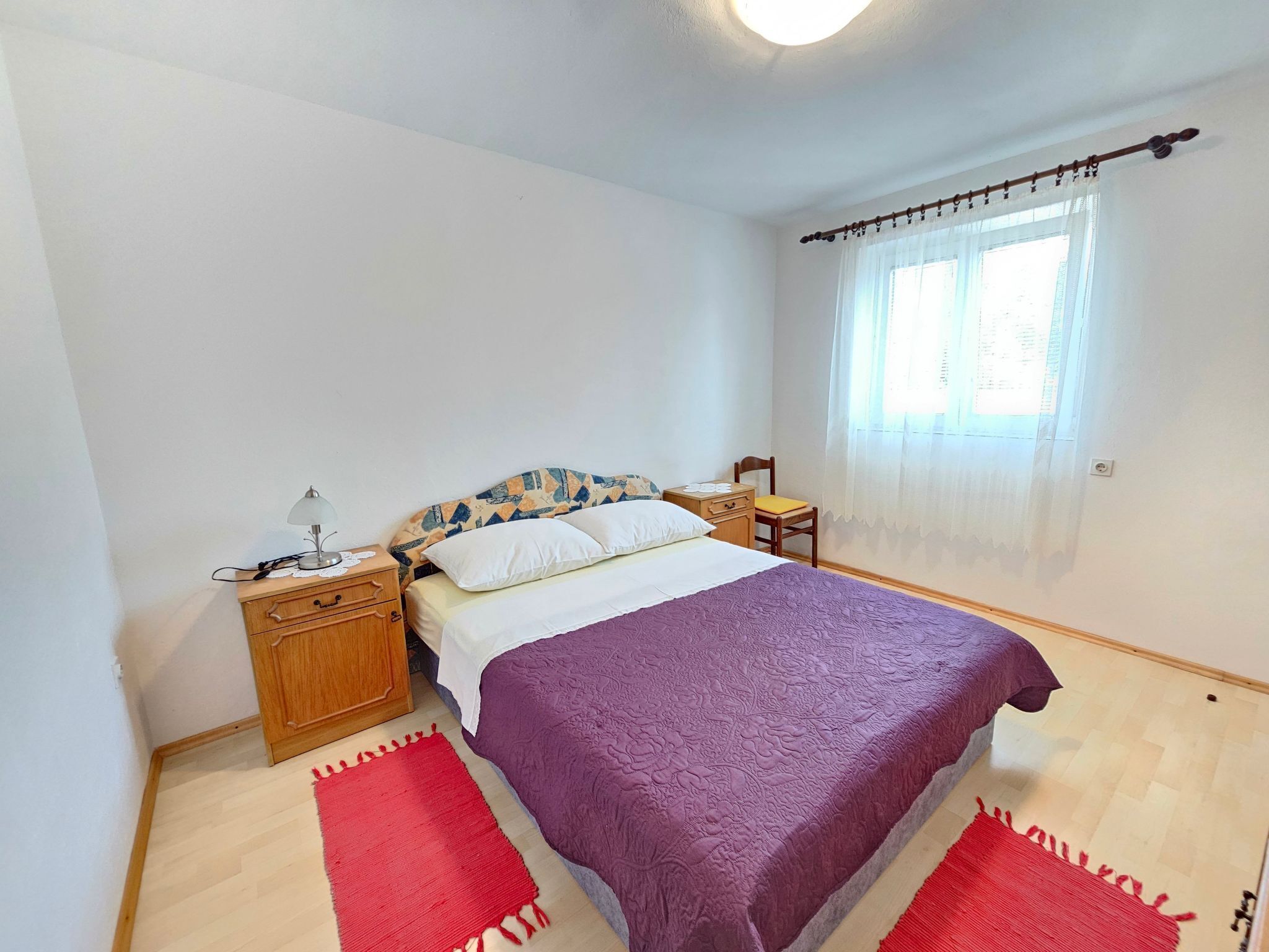 Ein Schlafzimmer mit Bett, Nachttisch, Lampe, Fenster mit Vorhängen und einem roten Teppich.