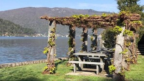 A wooden pergola with vines, a picnic table, and a grassy area by a lake with mountains in the background. - Exklusives Apartment, am Luganersee, mit Außenpool by Interhome (MAROGGIA)