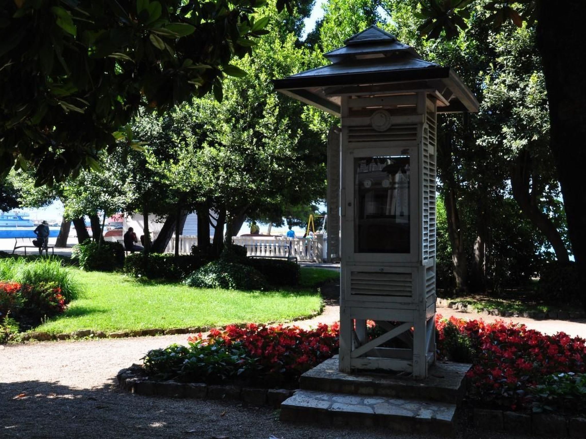 Un pabellón en un parque con vista al agua y personas caminando al fondo.