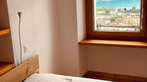 A bedroom with a bed, a wooden headboard, a window with a view of a mountainous landscape and a town, and a bedside lamp. - Ai Ronchi by Interhome (Gordola)