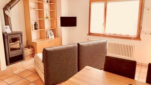 A cozy room with a fireplace, a TV, a dining table, and a window with blinds. - Ai Ronchi by Interhome (Gordola)