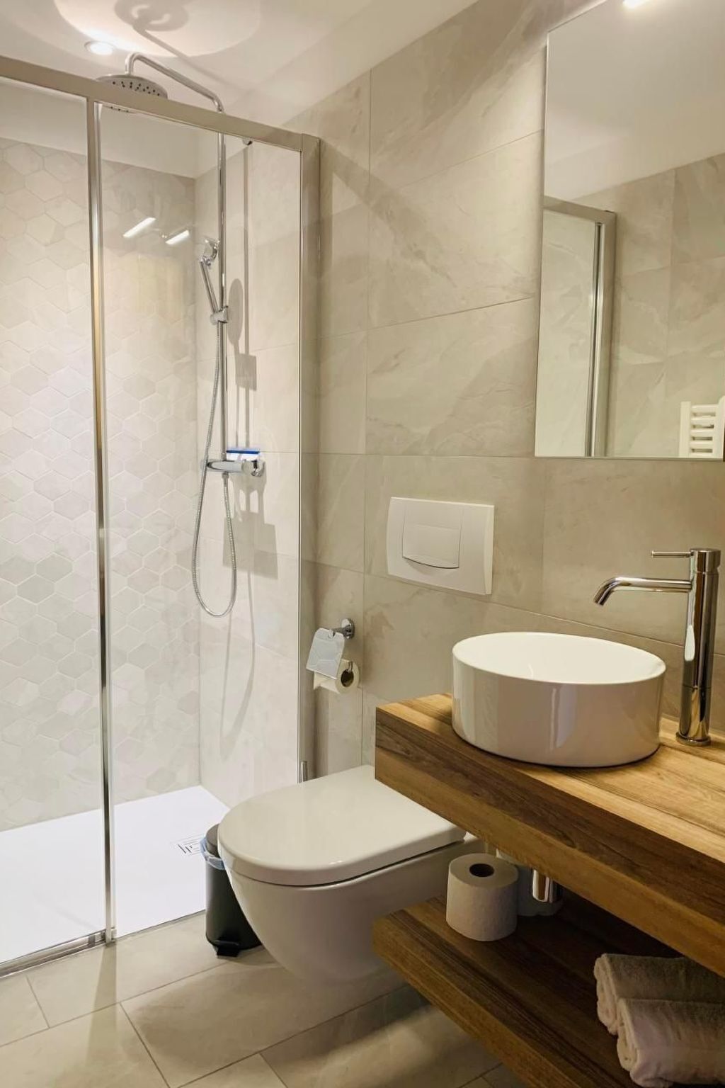 Een moderne badkamer met een douchecabine van glas, een wit toilet, een houten wastafelmeubel met een ronde wastafel en een spiegel boven de wastafel.