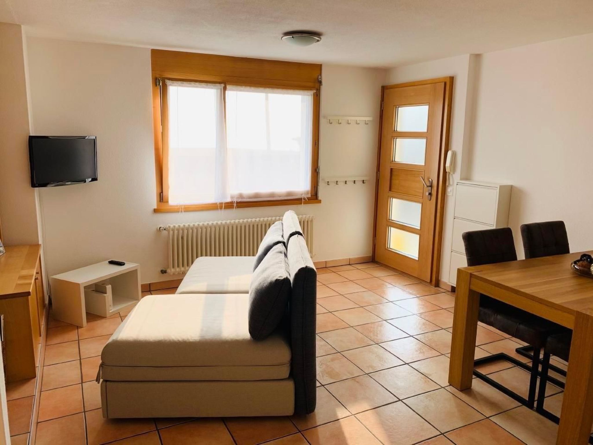 Een ruime, goed verlichte kamer met een bank, een televisie, een eettafel en een raam met gordijnen.